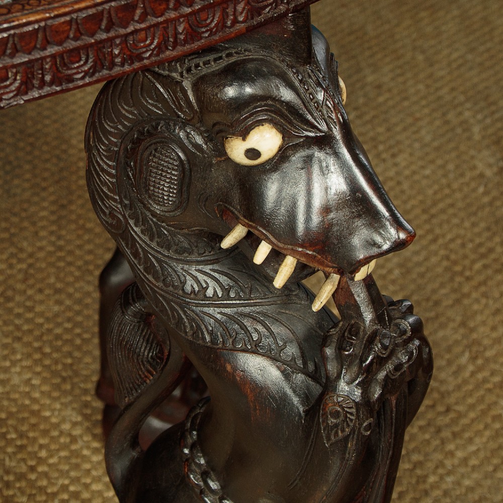 An oval dark hardwood Anglo-Indian coffee table featuring intricate top carvings and four legs sculpted as mythological beasts with inset white teeth. View VII