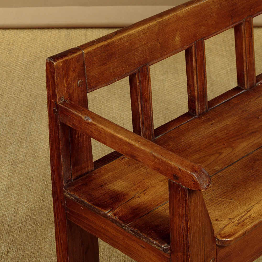 A small antique warm brown oak hall bench featuring a slatted open backrest, straight armrests, and a solid plank seat. View VI