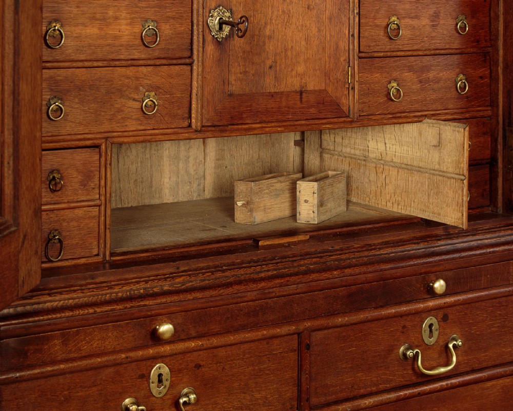 An early 18th-century oak cabinet on chest featuring twin panelled doors opening to internal drawers and pigeonholes above a chest of drawers with brass handles. View X