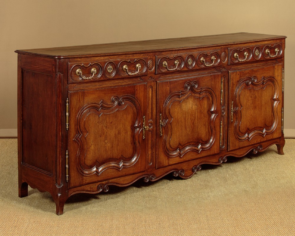 An 18th-century French provincial dark oak dresser base featuring three carved panel doors, three upper drawers with brass handles, and a shaped apron on short scrolled feet.