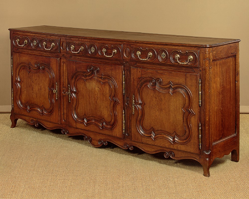 An 18th-century French provincial dark oak dresser base featuring three carved panel doors, three upper drawers with brass handles, and a shaped apron on short scrolled feet. View III