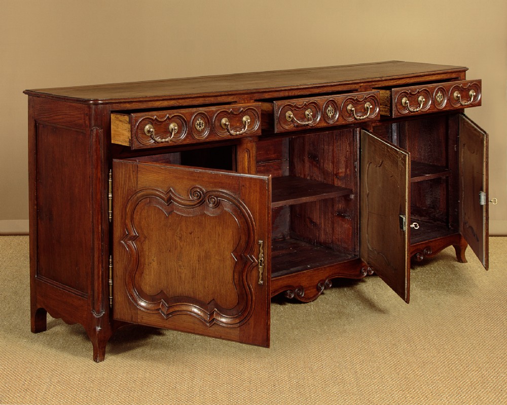 An 18th-century French provincial dark oak dresser base featuring three carved panel doors, three upper drawers with brass handles, and a shaped apron on short scrolled feet. View IV