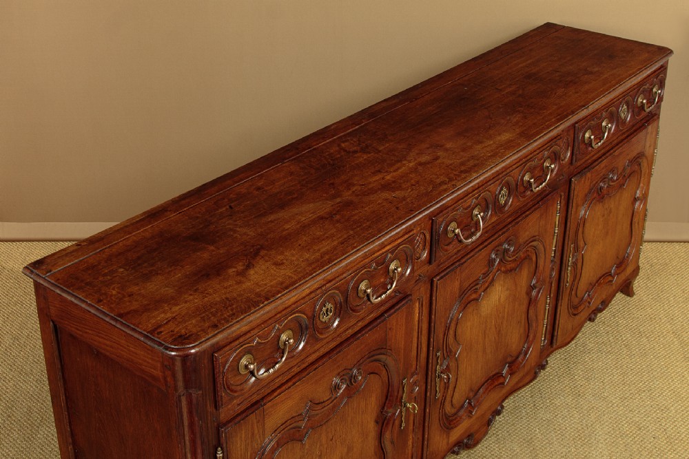 An 18th-century French provincial dark oak dresser base featuring three carved panel doors, three upper drawers with brass handles, and a shaped apron on short scrolled feet. View V
