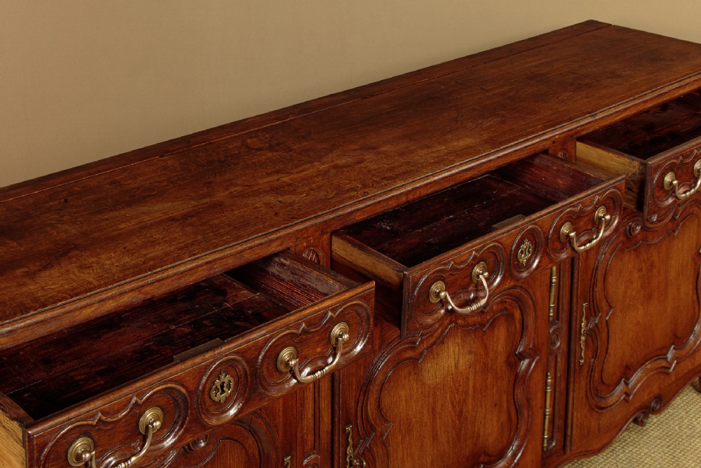 An 18th-century French provincial dark oak dresser base featuring three carved panel doors, three upper drawers with brass handles, and a shaped apron on short scrolled feet. View VI