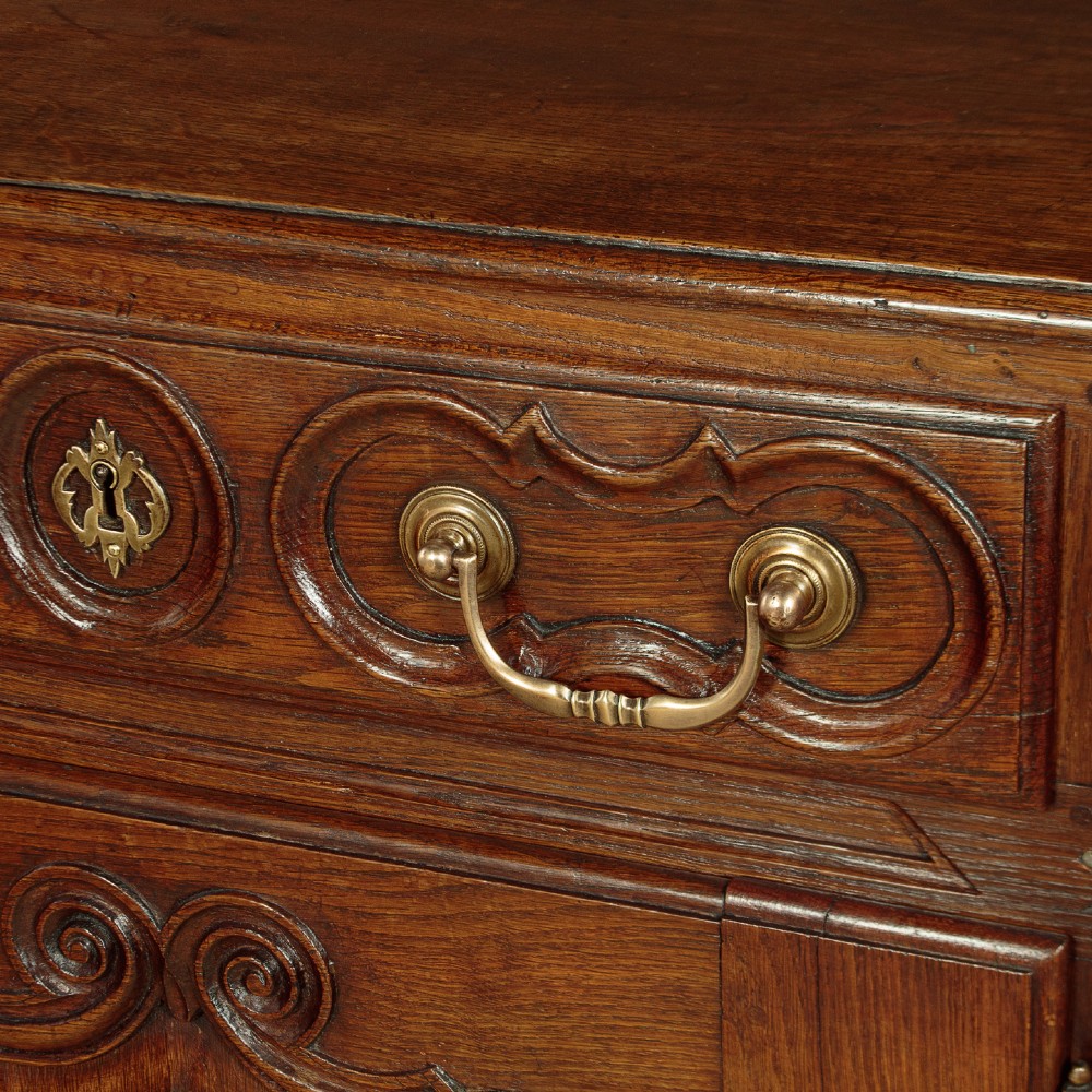 An 18th-century French provincial dark oak dresser base featuring three carved panel doors, three upper drawers with brass handles, and a shaped apron on short scrolled feet. View VIII