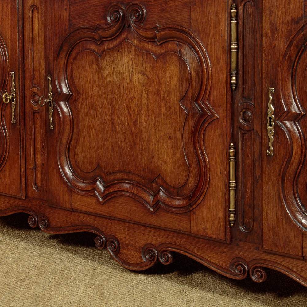 An 18th-century French provincial dark oak dresser base featuring three carved panel doors, three upper drawers with brass handles, and a shaped apron on short scrolled feet. View X
