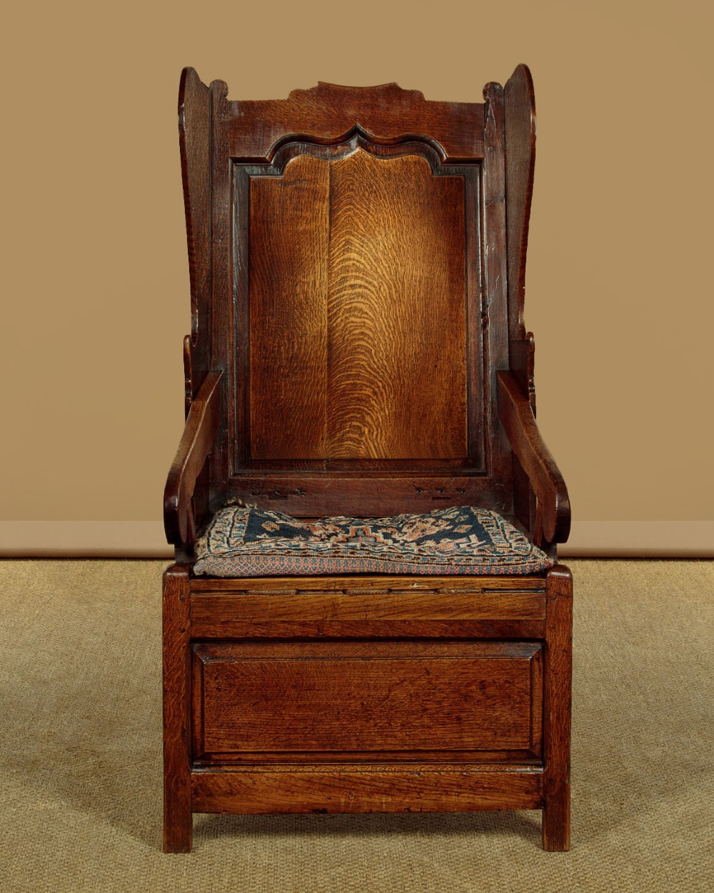 A late 18th-century dark oak lambing chair featuring high winged sides, a panelled back, a patterned seat cushion, and a drawer in the base.
