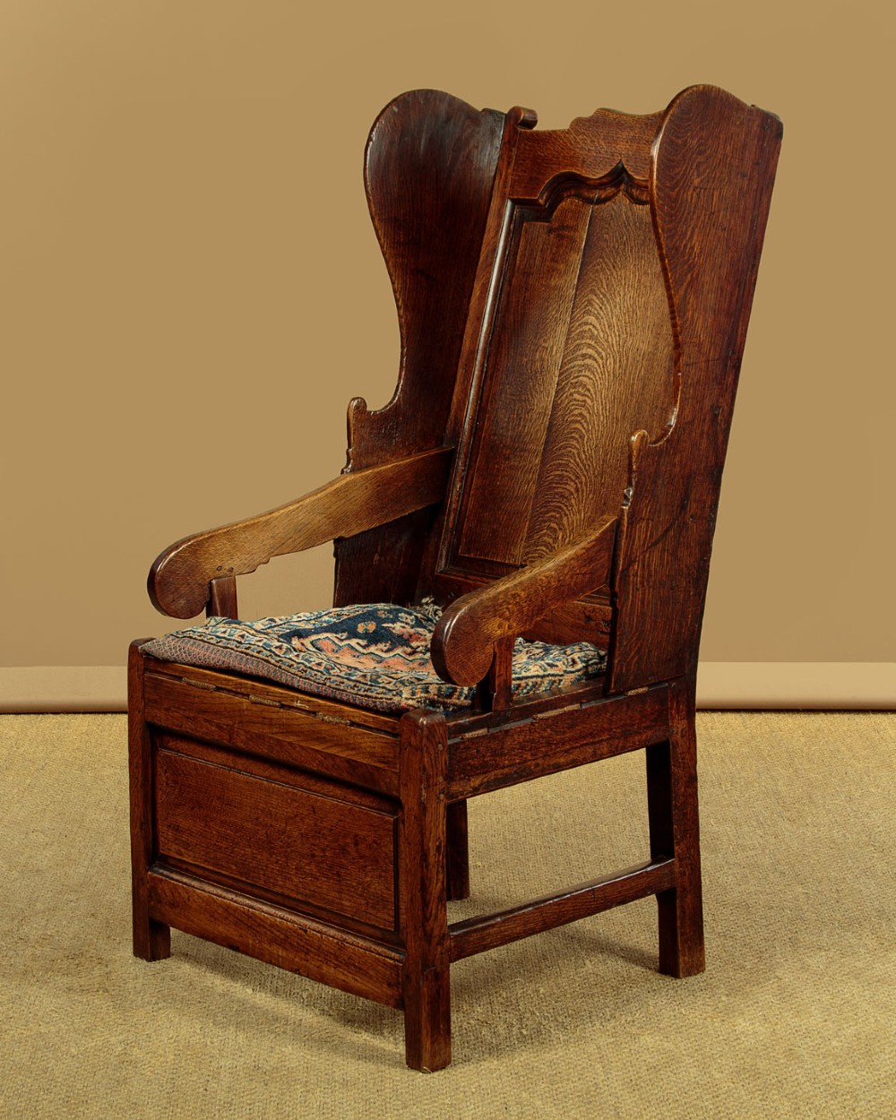 A late 18th-century dark oak lambing chair featuring high winged sides, a panelled back, a patterned seat cushion, and a drawer in the base. View III