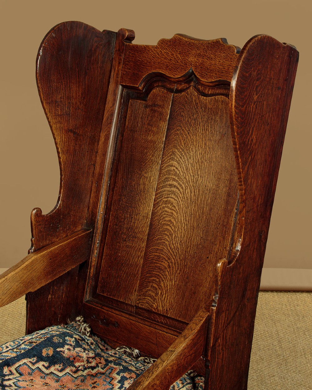 A late 18th-century dark oak lambing chair featuring high winged sides, a panelled back, a patterned seat cushion, and a drawer in the base. View V