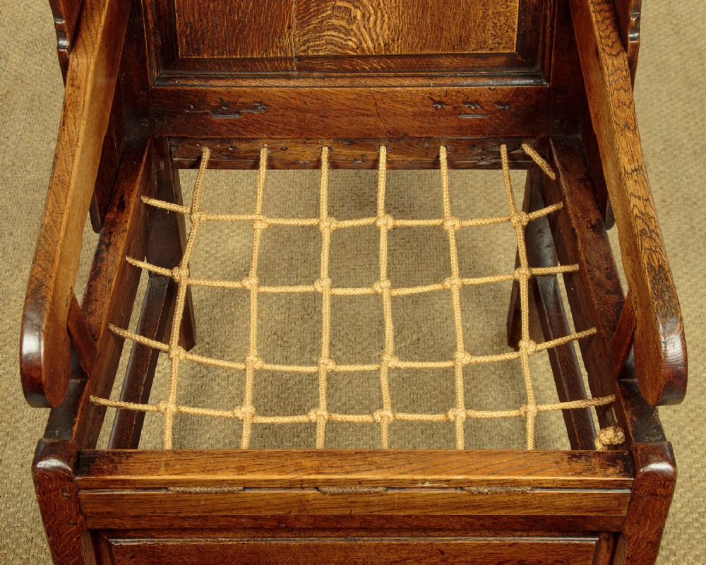 A late 18th-century dark oak lambing chair featuring high winged sides, a panelled back, a patterned seat cushion, and a drawer in the base. View X