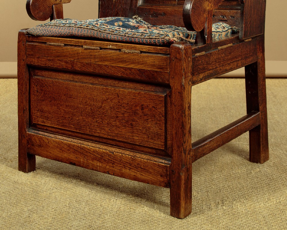 A late 18th-century dark oak lambing chair featuring high winged sides, a panelled back, a patterned seat cushion, and a drawer in the base. View XI