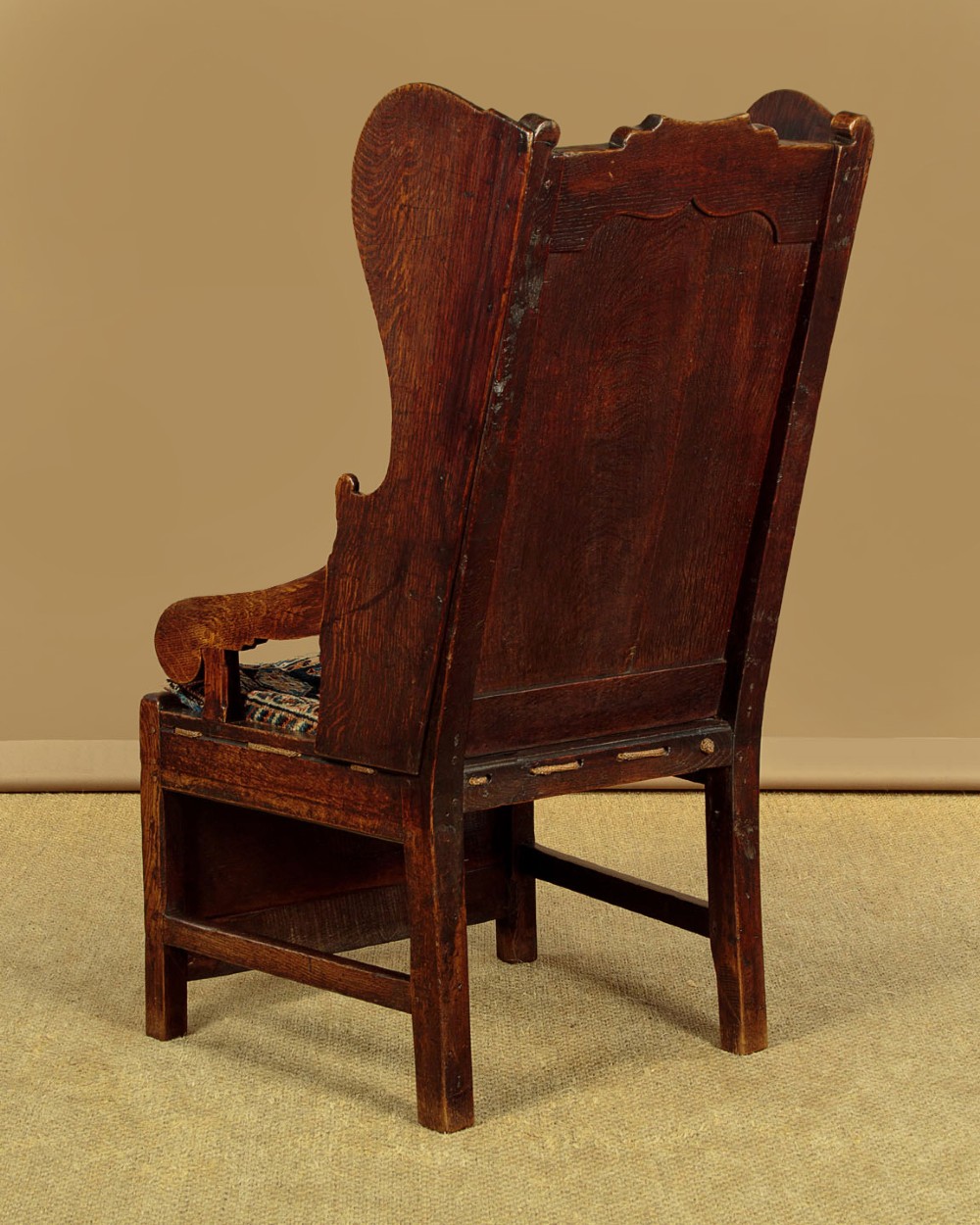 A late 18th-century dark oak lambing chair featuring high winged sides, a panelled back, a patterned seat cushion, and a drawer in the base. View XII