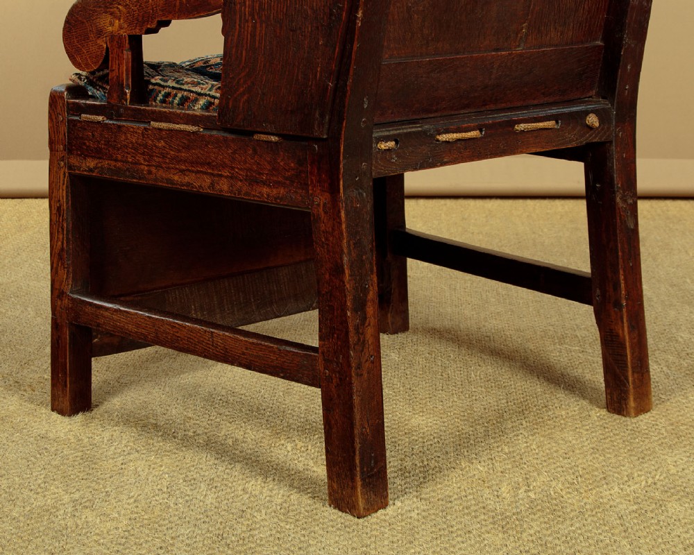A late 18th-century dark oak lambing chair featuring high winged sides, a panelled back, a patterned seat cushion, and a drawer in the base. View XIII