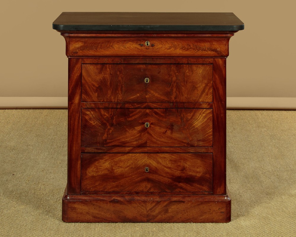 A nineteenth-century flame mahogany commode chest with four drawers, brass escutcheons, a dark slate top surface, and a plinth base.