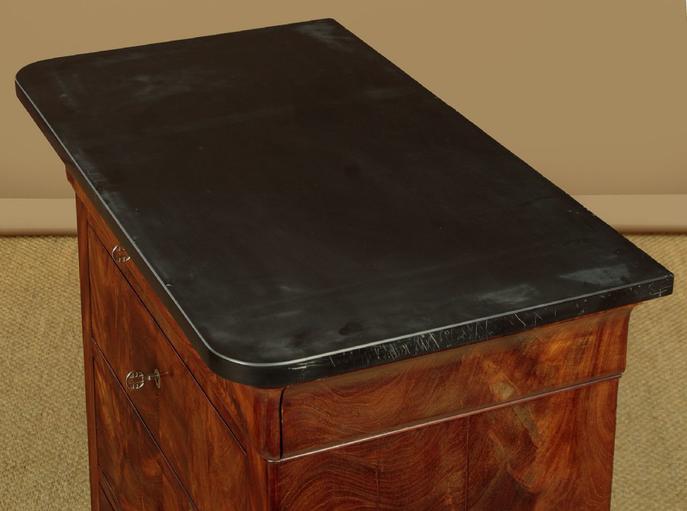 A nineteenth-century flame mahogany commode chest with four drawers, brass escutcheons, a dark slate top surface, and a plinth base. View V