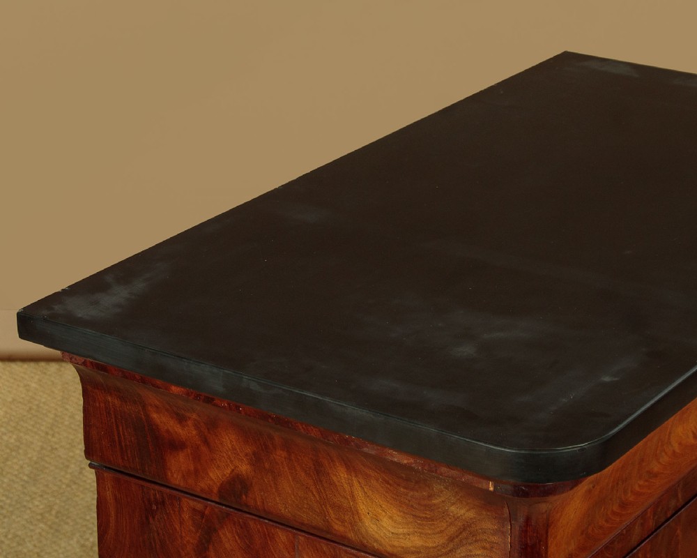 A nineteenth-century flame mahogany commode chest with four drawers, brass escutcheons, a dark slate top surface, and a plinth base. View VI