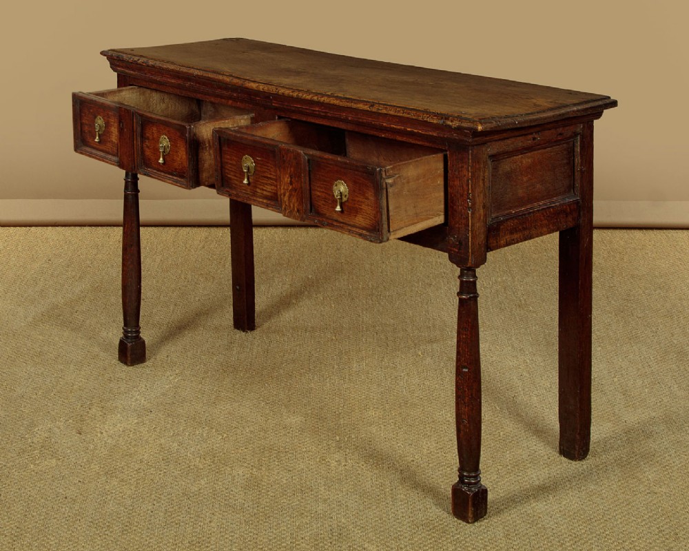 An 18th-century dark oak dresser base featuring geometric moulded drawer fronts, brass drop handles, and turned front legs. View V