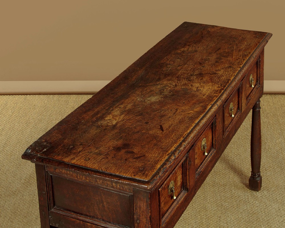 An 18th-century dark oak dresser base featuring geometric moulded drawer fronts, brass drop handles, and turned front legs. View VI