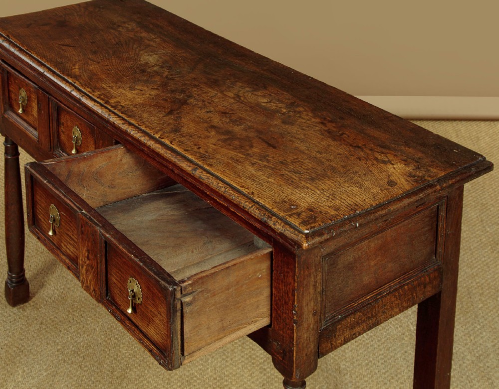 An 18th-century dark oak dresser base featuring geometric moulded drawer fronts, brass drop handles, and turned front legs. View VII