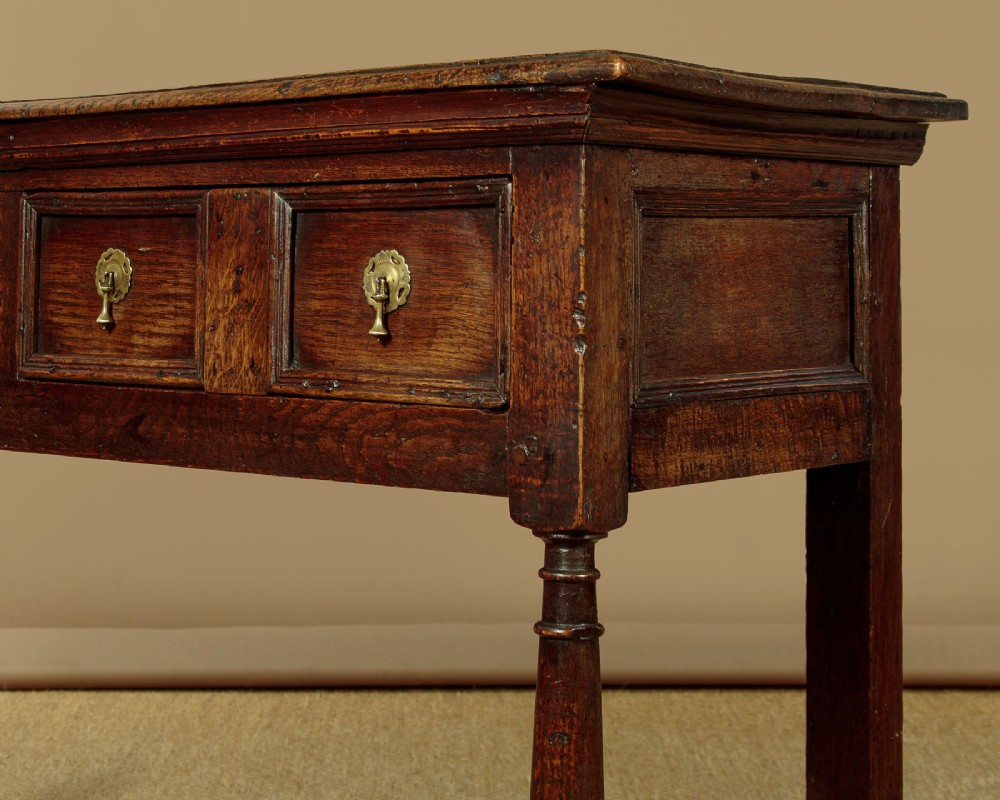 An 18th-century dark oak dresser base featuring geometric moulded drawer fronts, brass drop handles, and turned front legs. View IX