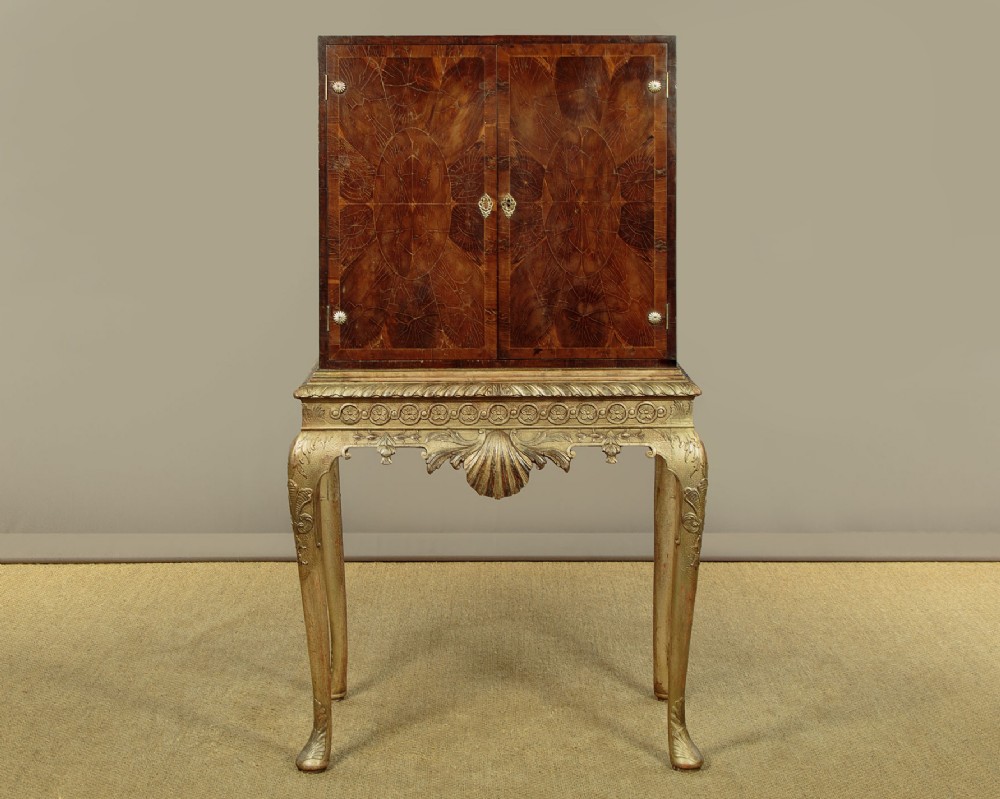 A small veneered brown wood cabinet featuring decorative door pulls, mounted atop a carved silver gilt stand with cabriole legs and a central shell motif.