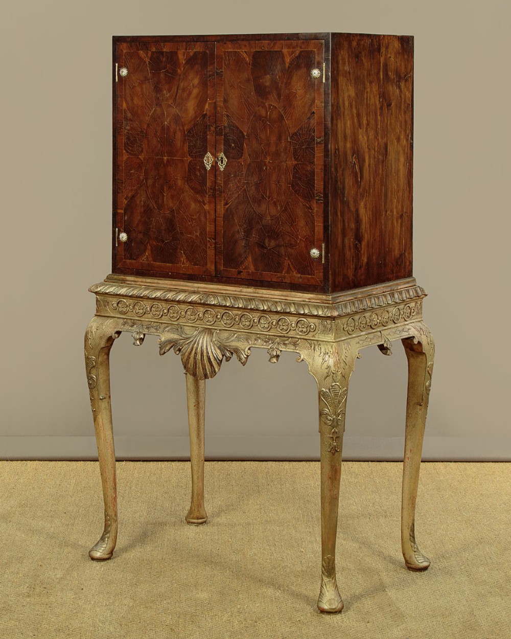 A small veneered brown wood cabinet featuring decorative door pulls, mounted atop a carved silver gilt stand with cabriole legs and a central shell motif. View IV