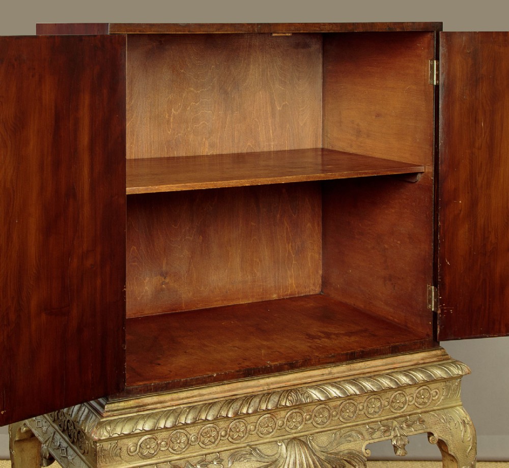 A small veneered brown wood cabinet featuring decorative door pulls, mounted atop a carved silver gilt stand with cabriole legs and a central shell motif. View VIII