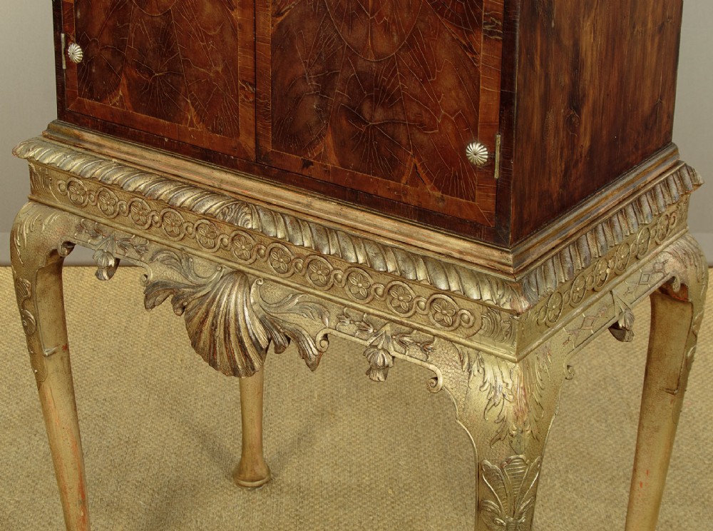 A small veneered brown wood cabinet featuring decorative door pulls, mounted atop a carved silver gilt stand with cabriole legs and a central shell motif. View X