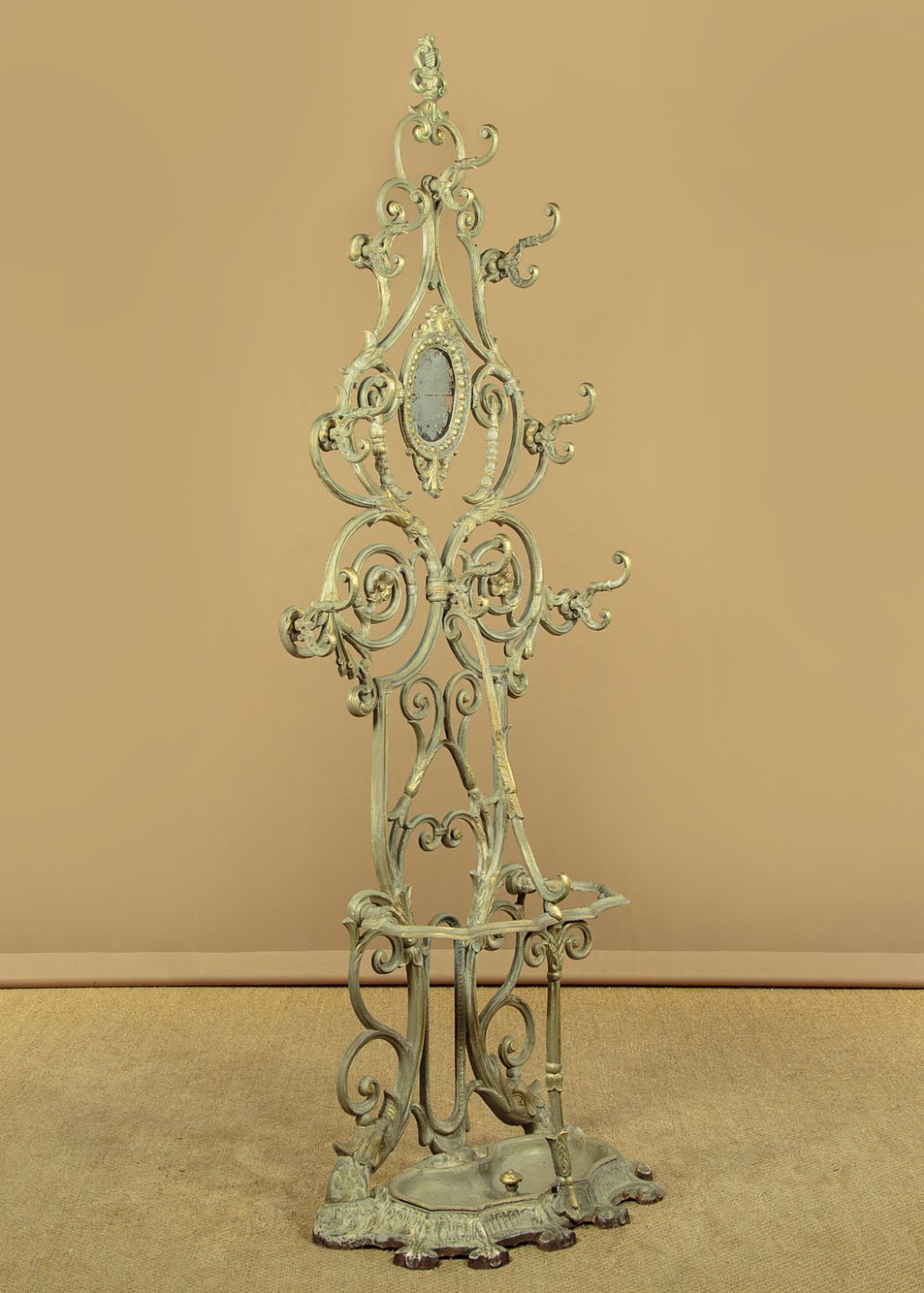 An ornate Victorian painted cast iron hall stand featuring scrollwork motifs, multiple coat hooks, a central oval mirror, and an umbrella drip tray base. View III