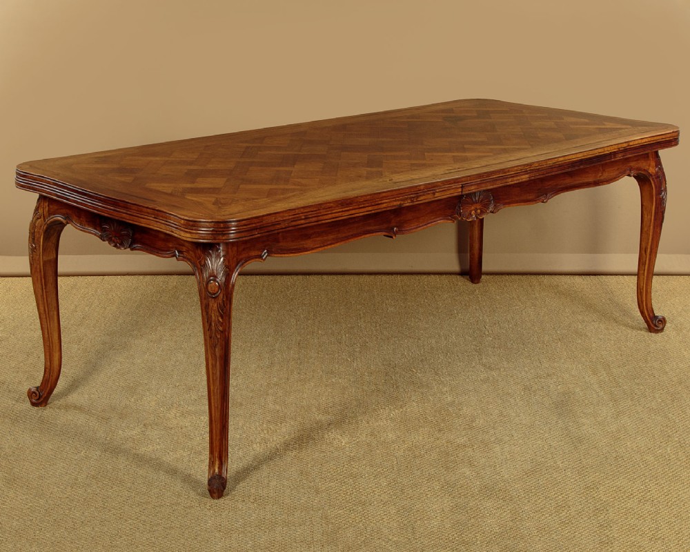 A rectangular French revival walnut dining table featuring a parquetry inlaid top, carved apron, and slender cabriole legs.