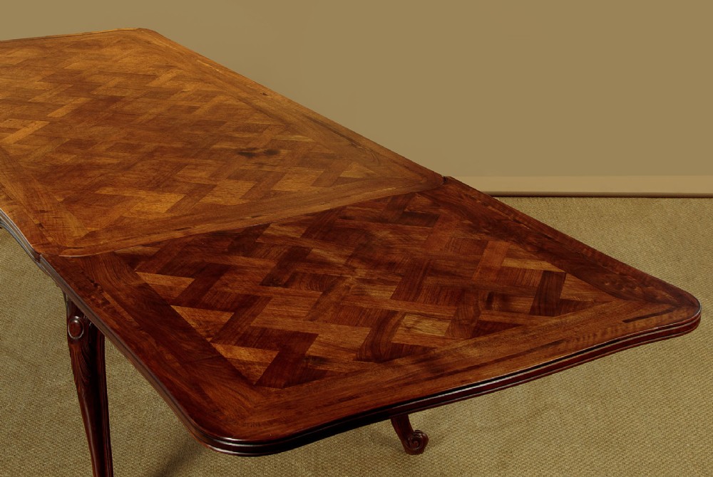 A rectangular French revival walnut dining table featuring a parquetry inlaid top, carved apron, and slender cabriole legs. View VII