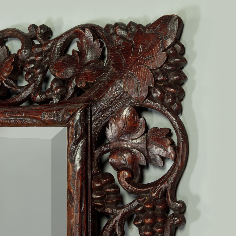 A rectangular dark oak wall mirror featuring intricate openwork carvings of grapevines and leaves framing a bevelled glass plate. View V