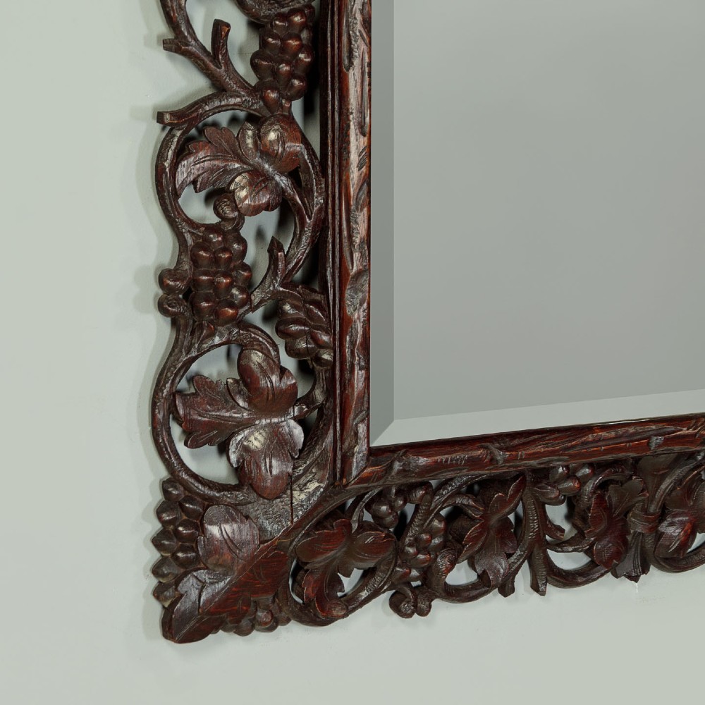 A rectangular dark oak wall mirror featuring intricate openwork carvings of grapevines and leaves framing a bevelled glass plate. View VII