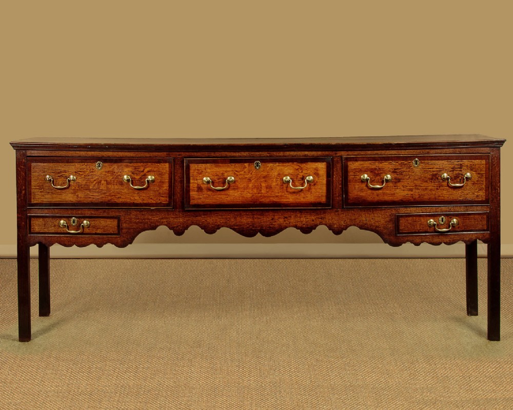 A long Georgian oak dresser base featuring five drawers with brass swan-neck handles, crossbanded drawer borders, a scalloped apron, and straight legs.