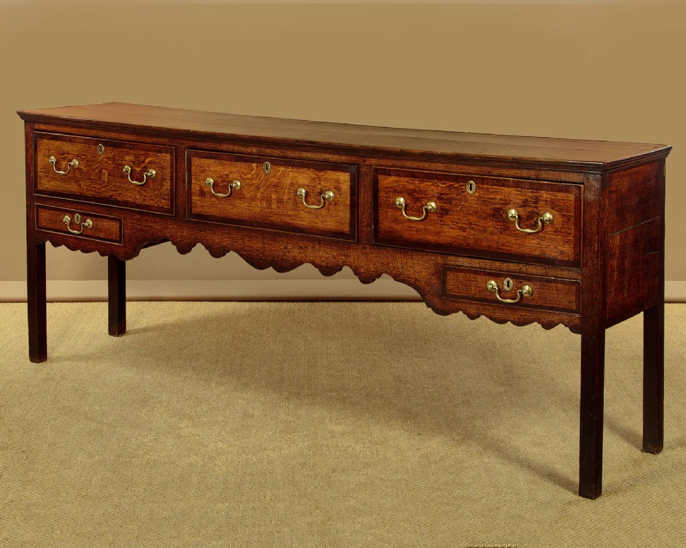 A long Georgian oak dresser base featuring five drawers with brass swan-neck handles, crossbanded drawer borders, a scalloped apron, and straight legs. View IV