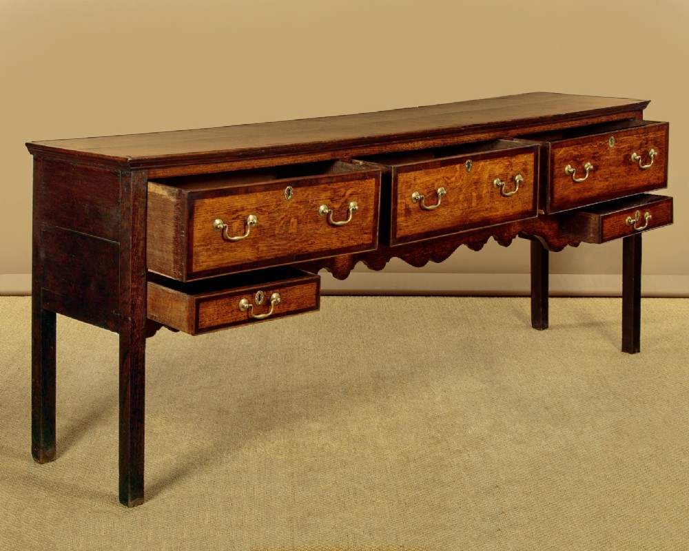 A long Georgian oak dresser base featuring five drawers with brass swan-neck handles, crossbanded drawer borders, a scalloped apron, and straight legs. View V