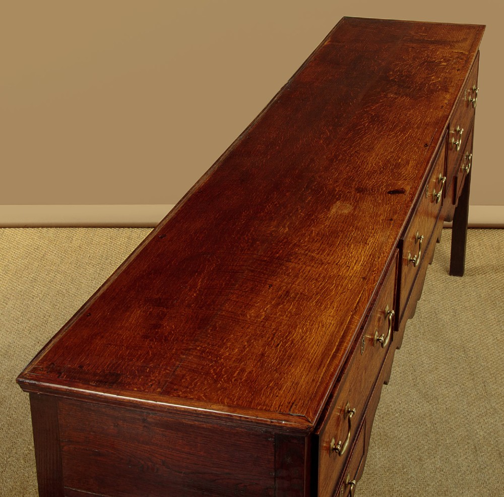 A long Georgian oak dresser base featuring five drawers with brass swan-neck handles, crossbanded drawer borders, a scalloped apron, and straight legs. View VI