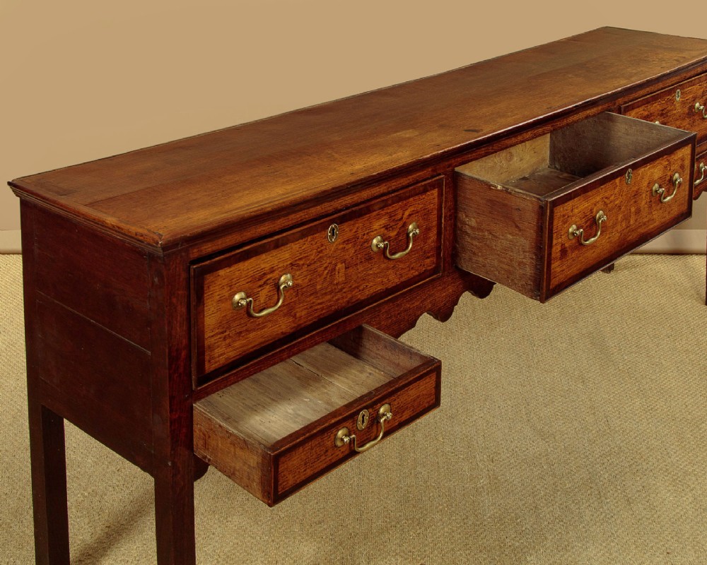 A long Georgian oak dresser base featuring five drawers with brass swan-neck handles, crossbanded drawer borders, a scalloped apron, and straight legs. View IX