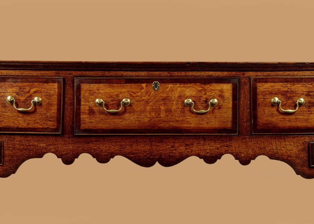 A long Georgian oak dresser base featuring five drawers with brass swan-neck handles, crossbanded drawer borders, a scalloped apron, and straight legs. View XI