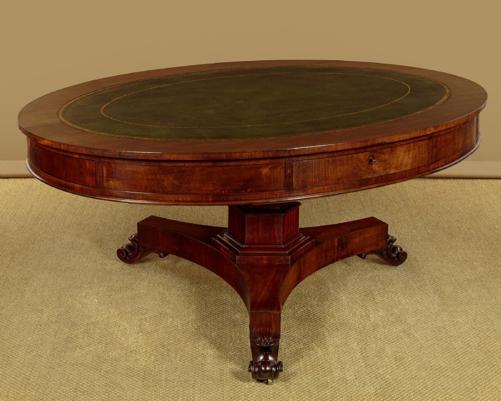 An oval Victorian mahogany library table featuring a green gilt-tooled leather top, frieze drawers, and a central pedestal with carved scroll feet on castors.