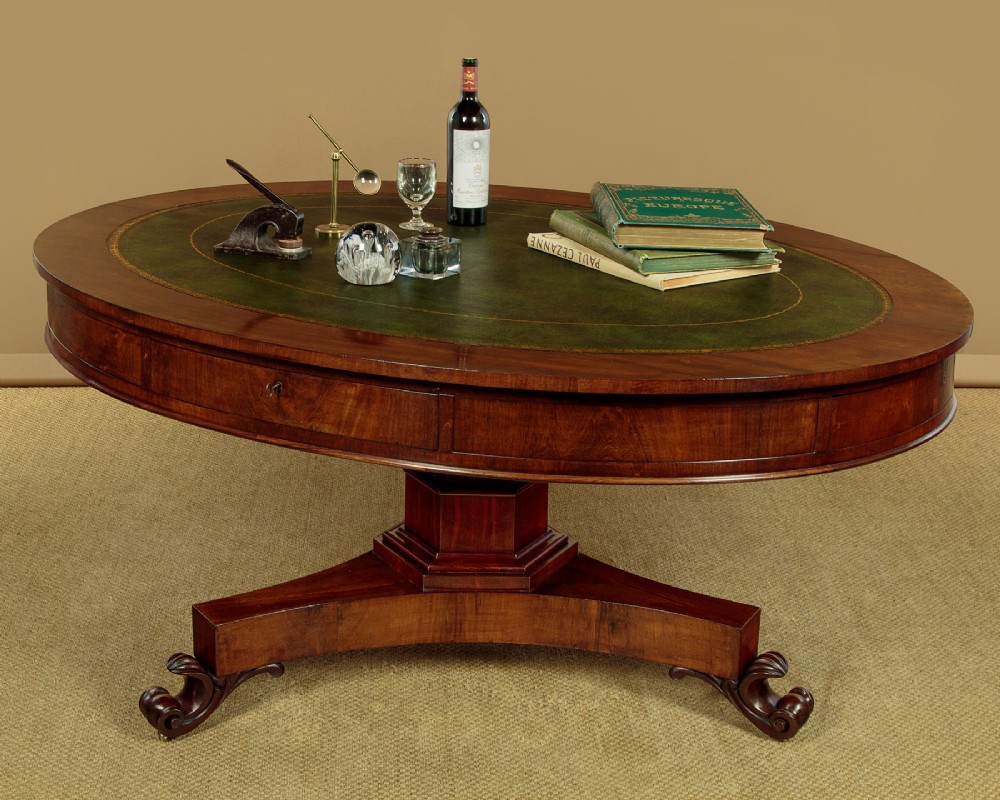 An oval Victorian mahogany library table featuring a green gilt-tooled leather top, frieze drawers, and a central pedestal with carved scroll feet on castors. View II