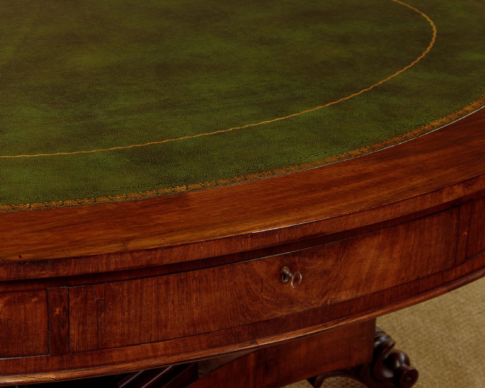 An oval Victorian mahogany library table featuring a green gilt-tooled leather top, frieze drawers, and a central pedestal with carved scroll feet on castors. View VII