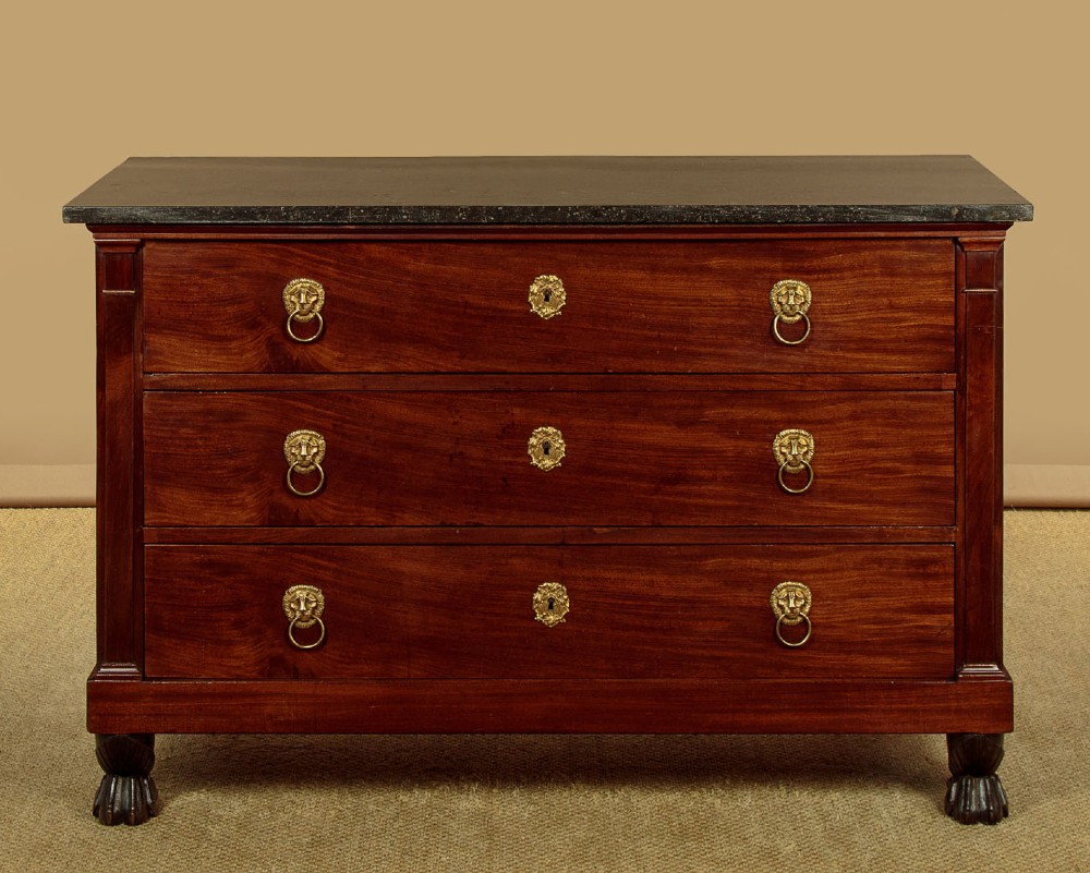 A French Empire mahogany commode featuring three long drawers, brass lion mask ring handles, carved claw feet, and a dark grey marble top.