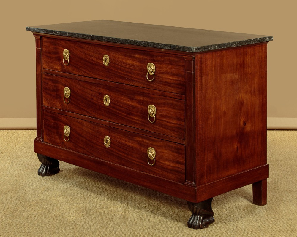 A French Empire mahogany commode featuring three long drawers, brass lion mask ring handles, carved claw feet, and a dark grey marble top. View IV