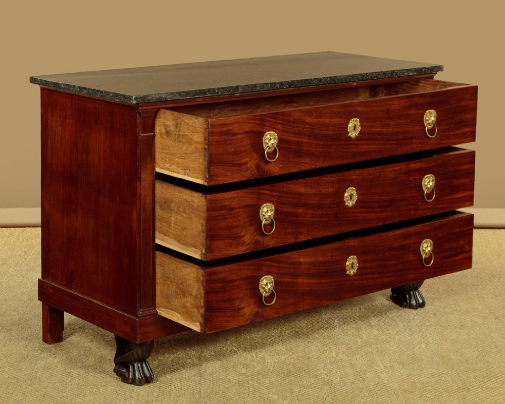 A French Empire mahogany commode featuring three long drawers, brass lion mask ring handles, carved claw feet, and a dark grey marble top. View V