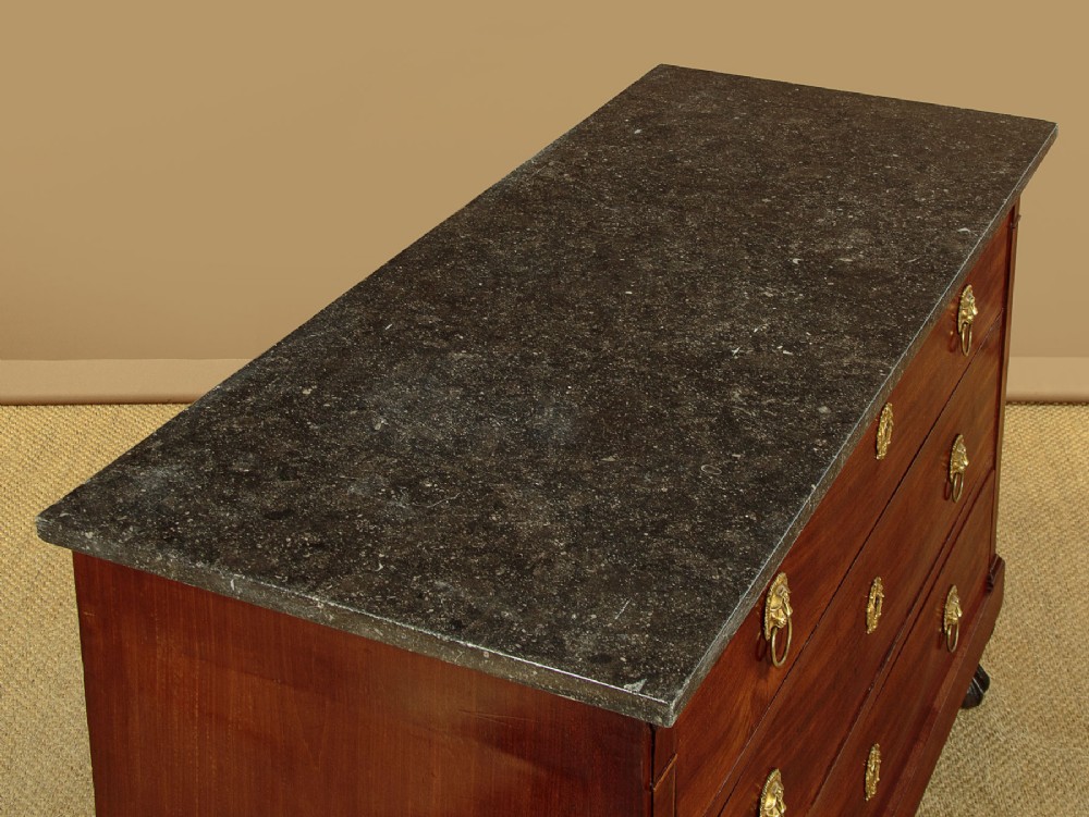 A French Empire mahogany commode featuring three long drawers, brass lion mask ring handles, carved claw feet, and a dark grey marble top. View VI