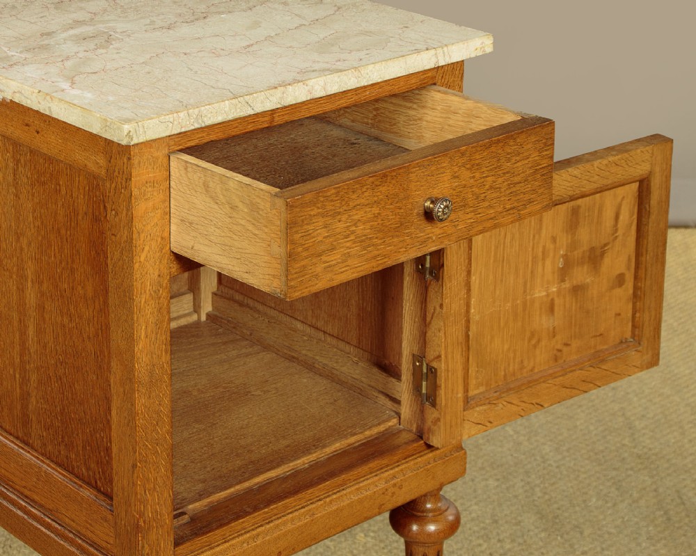 A pair of golden oak bedside cabinets featuring marble tops, single drawers, floral carved cupboard doors, and slim fluted turned legs. View X