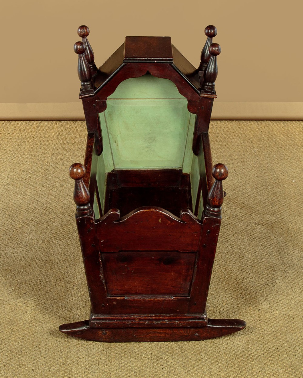 An antique dark brown wooden hooded rocking cradle featuring turned corner finials, a curved rocker base, and a pale green painted interior. View II