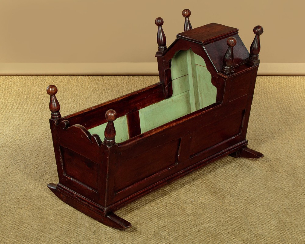 An antique dark brown wooden hooded rocking cradle featuring turned corner finials, a curved rocker base, and a pale green painted interior. View III