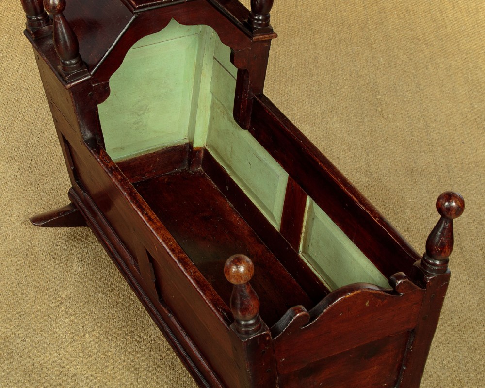 An antique dark brown wooden hooded rocking cradle featuring turned corner finials, a curved rocker base, and a pale green painted interior. View VI